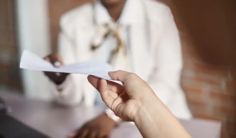 obrazek wyróżniający w artykule - Podanie o podwyżkę. Na zdjęciu widać dwie osoby, z których jedna przekazuje dokument drugiej. Na pierwszym planie, w prawej dolnej części zdjęcia, znajduje się ręka osoby trzymającej biały papierowy dokument. W tle, nieco rozmyta, jest druga osoba, która zdaje się przyjmować dokument. Ta osoba ma na sobie jasną marynarkę i bluzkę, a także elegancką apaszkę zawiązaną na szyi. Scena sugeruje formalne spotkanie, prawdopodobnie w biurze lub urzędzie, gdzie przekazywane są ważne dokumenty.
