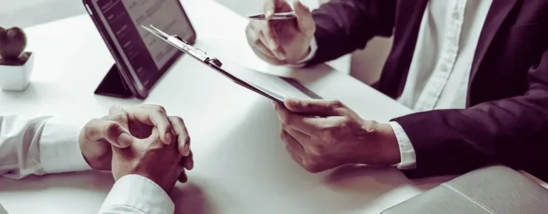 obrazek wyróżniający w artykule - słabe strony podczas rozmowy kwalifikacyjnej. Na obrazie widzimy fragment sceny, która najprawdopodobniej przedstawia rozmowę kwalifikacyjną lub spotkanie biznesowe. Dwie osoby siedzą przy biurku. Osoba po lewej stronie ma złożone dłonie i jest ubrana w białą koszulę, natomiast osoba po prawej stronie, ubrana w ciemny garnitur, trzyma w ręku długopis i tabliczkę z dokumentami. Na biurku znajduje się również tablet oraz zamknięty notes lub folder. Ujęcie jest zbliżone na ręce obu osób oraz na dokumenty, co sugeruje, że mogą omawiać istotne szczegóły lub podpisywać umowę.