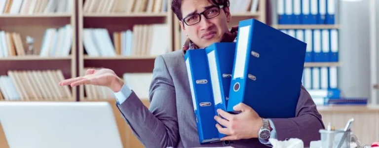 obrazek wyróżniający w artykule - czy można zwolnić pracownika na l4. Na zdjęciu widoczny jest mężczyzna w biurze, ubrany w elegancki garnitur. Trzyma on trzy niebieskie segregatory, wyglądające na ciężkie, co sugeruje dużą ilość dokumentów do przejrzenia lub pracy do wykonania. Jego wyraz twarzy wyraża zakłopotanie lub frustrację, a gest prawej ręki, z dłonią uniesioną i otwartą, sugeruje, że nie wie, co zrobić lub jest zdezorientowany. W tle widoczne są półki z książkami i dokumentami, co podkreśla biurowe otoczenie. Wygląda na to, że mężczyzna ma do czynienia z nadmiarem obowiązków lub trudnościami związanymi z zarządzaniem dokumentacją.