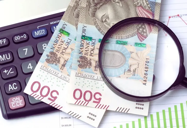 obrazek wyróżniający w artykule - obniżenie wpłaty na PFRON. Zdjęcie przedstawia kilka polskich banknotów o nominale 500 zł, które leżą na powierzchni wraz z kalkulatorem i szkłem powiększającym. Szkło powiększające jest umieszczone nad jednym z banknotów, co podkreśla szczegóły widoczne na banknocie. Kalkulator znajduje się po lewej stronie zdjęcia, a pod banknotami widać fragmenty wykresów na kartce papieru. Kolorystyka jest stonowana, dominują odcienie niebieskiego, zielonego i czerwonego. Cała scena sugeruje kontekst finansowy lub związany z analizą kosztów.