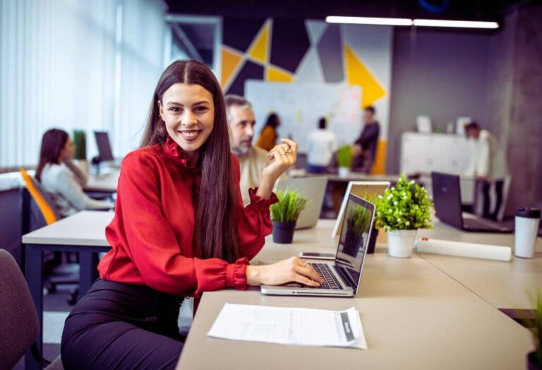 obrazek wyróżniający w artykule - outsourcing pracowniczy. Na zdjęciu widzimy młodą kobietę siedzącą przy biurku w biurze typu open space. Uśmiecha się szeroko, patrząc w stronę aparatu. Ma długie, proste, ciemne włosy i ubrana jest w czerwoną bluzkę z długimi rękawami i ozdobnymi mankietami. Pracuje na laptopie, a na biurku przed nią leży dokument. W tle widać nowoczesne biuro z kilkoma osobami pracującymi przy innych stanowiskach. Ściany są ozdobione geometrycznymi wzorami w odcieniach szarości, bieli i żółtego. Na biurkach znajdują się zielone rośliny doniczkowe, które dodają świeżości do przestrzeni. Atmosfera wydaje się profesjonalna, ale jednocześnie przyjazna i swobodna, sugerująca współczesne środowisko pracy.