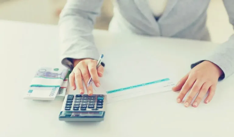 obrazek wyróżniający w artykule - dofinansowanie na pracownika niepełnosprawnego. Na obrazie widzimy osobę siedzącą przy białym stole, ubranej w elegancki, szary garnitur. Osoba wykonuje obliczenia za pomocą kalkulatora, jednocześnie trzymając długopis w ręce. Na stole, obok kalkulatora, znajduje się plik banknotów, a także papierowy dokument, który prawdopodobnie może być czekiem lub formularzem. Obraz sugeruje, że osoba ta zajmuje się finansami lub pracą biurową, związaną z rozliczeniami. Stylizowany jest na profesjonalne środowisko pracy, z naciskiem na kwestie finansowe.