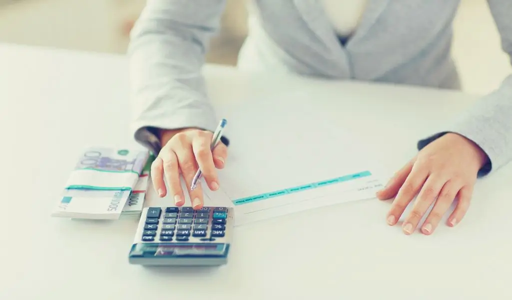 Przewodnik dla pracodawcy, o jakie dofinansowanie na pracownika niepełnosprawnego może się ubiegać 1 obrazek wyróżniający w artykule - dofinansowanie na pracownika niepełnosprawnego. Na obrazie widzimy osobę siedzącą przy białym stole, ubranej w elegancki, szary garnitur. Osoba wykonuje obliczenia za pomocą kalkulatora, jednocześnie trzymając długopis w ręce. Na stole, obok kalkulatora, znajduje się plik banknotów, a także papierowy dokument, który prawdopodobnie może być czekiem lub formularzem. Obraz sugeruje, że osoba ta zajmuje się finansami lub pracą biurową, związaną z rozliczeniami. Stylizowany jest na profesjonalne środowisko pracy, z naciskiem na kwestie finansowe.