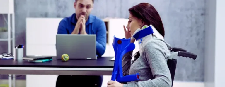 obrazek wyróżniający w artykule - uprawnienia pracowników z orzeczeniem. Na zdjęciu widzimy kobietę w kołnierzu ortopedycznym, siedzącą na wózku inwalidzkim. W jednej ręce trzyma niebieskie kule ortopedyczne, co sugeruje, że jest w trakcie leczenia lub rekonwalescencji po urazie. Jej wyraz twarzy wskazuje na zmęczenie lub niepokój, a ręką lekko dotyka twarzy, co może sugerować stres lub frustrację. Po drugiej stronie biurka siedzi mężczyzna przy komputerze, ubrany w niebieską koszulę. Ma złożone ręce i patrzy na kobietę z zamyśleniem, co może sugerować, że prowadzą rozmowę dotyczącą jej sytuacji zdrowotnej lub zawodowej. Cała scena wydaje się być formalna, prawdopodobnie odbywa się w kontekście pracy lub wsparcia zawodowego.