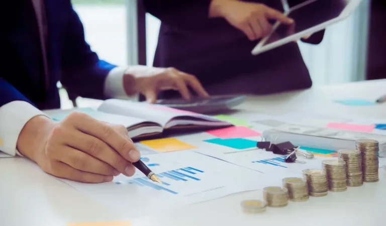 zdjęcie do artykułu - warunki otrzymania dofinansowania. Na obrazku widzimy biurową scenę pracy, w której główną uwagę przyciąga ręka mężczyzny trzymająca elegancki długopis nad dokumentem z wykresami. Obok leży otwarty notes oraz różnokolorowe karteczki samoprzylepne, które dodają dynamiki i sugerują planowanie lub burzę mózgów. W tle inna osoba trzyma tablet, co podkreśla użycie nowoczesnych technologii w analizie danych. Na pierwszym planie widnieją równo ułożone stosy monet, symbolizujące finanse, inwestycje lub budżetowanie. Obrazek tworzy atmosferę skupienia, profesjonalizmu i współpracy, budząc wrażenie zaangażowania w ważne decyzje biznesowe.